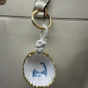 May include: A white seashell ornament with a gold-painted edge and a blue silhouette of Cape Cod painted on the interior. The shell hangs from a gold-toned metal ring by a thick, knotted white cord, attached to a textured off-white handbag.