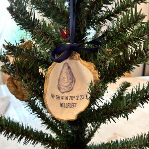 May include: A decorative oyster shell ornament featuring a hand-painted blue oyster illustration, gold-painted edges, and black text reading 41° 56' 4" N 70° 2' 23" W WELLFLEET. The shell hangs from a dark blue satin ribbon tied in a bow at the top.