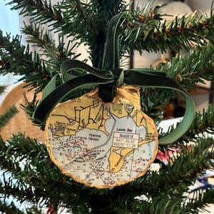 May include: A round seashell ornament featuring a nautical map of Hyannis Harbor and Lewis Bay, Massachusetts. The shell has a gold-painted edge and is suspended by a dark green velvet ribbon tied in a bow at the top of the shell.