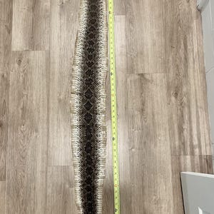 Puede incluir: Una larga piel de serpiente de cascabel mudada que presenta un patrón de diamantes de color marrón oscuro y crema a lo largo del centro. Una cinta métrica amarilla se coloca junto a la piel, indicando una longitud total de 132 cm.