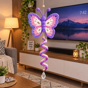 May include: A hanging crochet butterfly ornament featuring purple, pink, and white yarn. The butterfly is attached to a spiraling, multi-colored crochet tail that ends with a clear, faceted crystal drop, accented by small wooden beads.