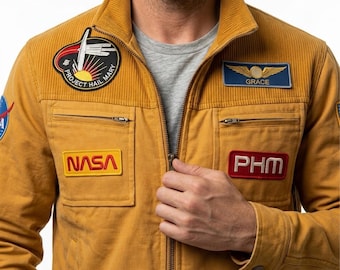 Puede incluir: Una chaqueta de color amarillo mostaza con cremallera, cuello acanalado y varios parches bordados. Los parches incluyen el emblema de la misión Project Hail Mary, un logotipo de la NASA, una insignia PHM y una etiqueta azul Grace con alas doradas.