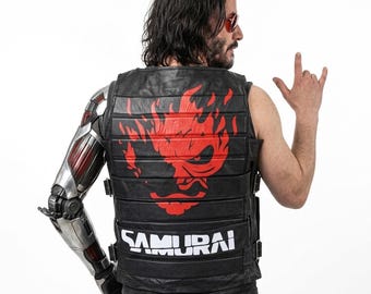 Puede incluir: Un chaleco de cuero negro con secciones acolchadas horizontales, un gran gráfico rojo de un rostro estilizado en la espalda y la palabra SAMURAI impresa en letras blancas en la parte inferior. El chaleco incluye correas con hebillas y un brazo robótico.