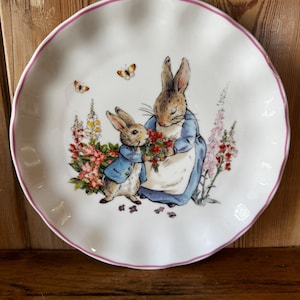 May include: A round white ceramic plate with a scalloped edge and a thin pink rim. The center features an illustration of two rabbits in blue jackets, one holding a bouquet of red flowers, surrounded by colorful garden flowers and two butterflies.