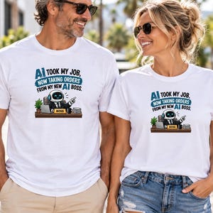Op de afbeelding: Een wit T-shirt met korte mouwen en een afbeelding van een robot die aan een bureau zit met een laptop en een bordje met de tekst BOSS. De tekst boven de robot luidt AI TOOK MY JOB, NOW TAKING ORDERS FROM MY NEW AI BOSS in blauwe letters.