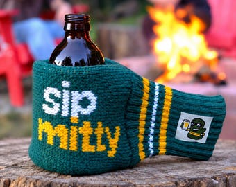 Puede incluir: Una funda de punto para botella en color verde oscuro con el texto "sip mitty" en blanco y amarillo en la parte frontal. La funda tiene rayas amarillas y blancas en el lateral y un pequeño parche cuadrado con un personaje de dibujos.
