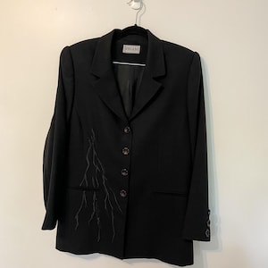 Peut inclure: Un blazer noir avec un revers cranté, quatre boutons sur le devant et des manches longues avec trois boutons à chaque poignet. Le vêtement présente un motif discret de foudre brodé sur le panneau avant droit et une étiquette de marque Priani.