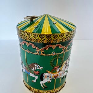 May include: A cylindrical metal tin featuring a green background with carousel horse illustrations, a gold and green striped conical lid, and a silver winding key on top. The design includes decorative gold scrollwork and festive wreaths.