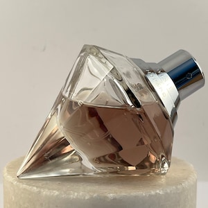 Könnte beinhalten: Eine durchsichtige Glas-Parfümflasche mit silbernem Verschluss. Die Flasche ist diamantförmig und enthält eine rosa Flüssigkeit. Die Flasche steht auf einer weißen Marmorfläche.