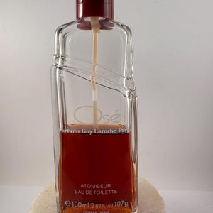 May include: A clear glass bottle of "Osé" perfume by Guy Laroche Paris. The bottle is filled with a brown liquid and has a red atomiser. The label reads "Atomiseur Eau de Toilette" and "e100 ml 3.89% vol 107g Cospar Paris".