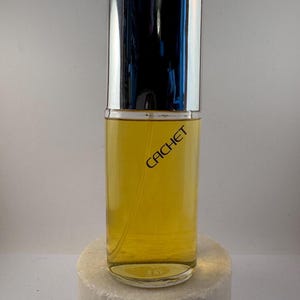 Könnte beinhalten: Eine durchsichtige Glasflasche mit Cachet-Parfum und einem silbernen Verschluss. Die Flasche ist mit einer gelben Flüssigkeit gefüllt und der Markenname ist in Schwarz auf der Vorderseite aufgedruckt.
