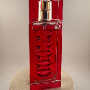 Puede incluir: Un frasco de perfume rojo rectangular con una tapa dorada. La botella tiene una serie de hendiduras curvas en la parte delantera. El frasco de perfume está sobre una superficie de piedra blanca, con un fondo rojo.