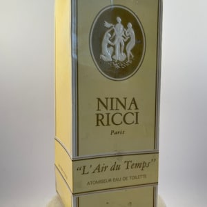Pode incluir: Uma caixa amarela com uma borda dourada e um círculo branco com um desenho de três figuras. O texto na caixa diz "Nina Ricci Paris" e "L'Air du Temps" Atomiseur Eau de Toilette.