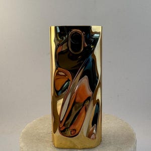 May include: A gold metal vase with a unique, wavy, abstract design. The vase is made of polished metal and has a reflective surface.