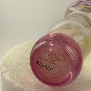 Puede incluir: Un frasco de perfume de vidrio transparente con una base rosa y una parte superior redondeada. El frasco tiene el texto "VERSACE WOMAN EAU DE PARFUM" y "1.7 US FL.OZ." impreso en la parte inferior. El frasco est&aacute; sobre una superficie blanca.