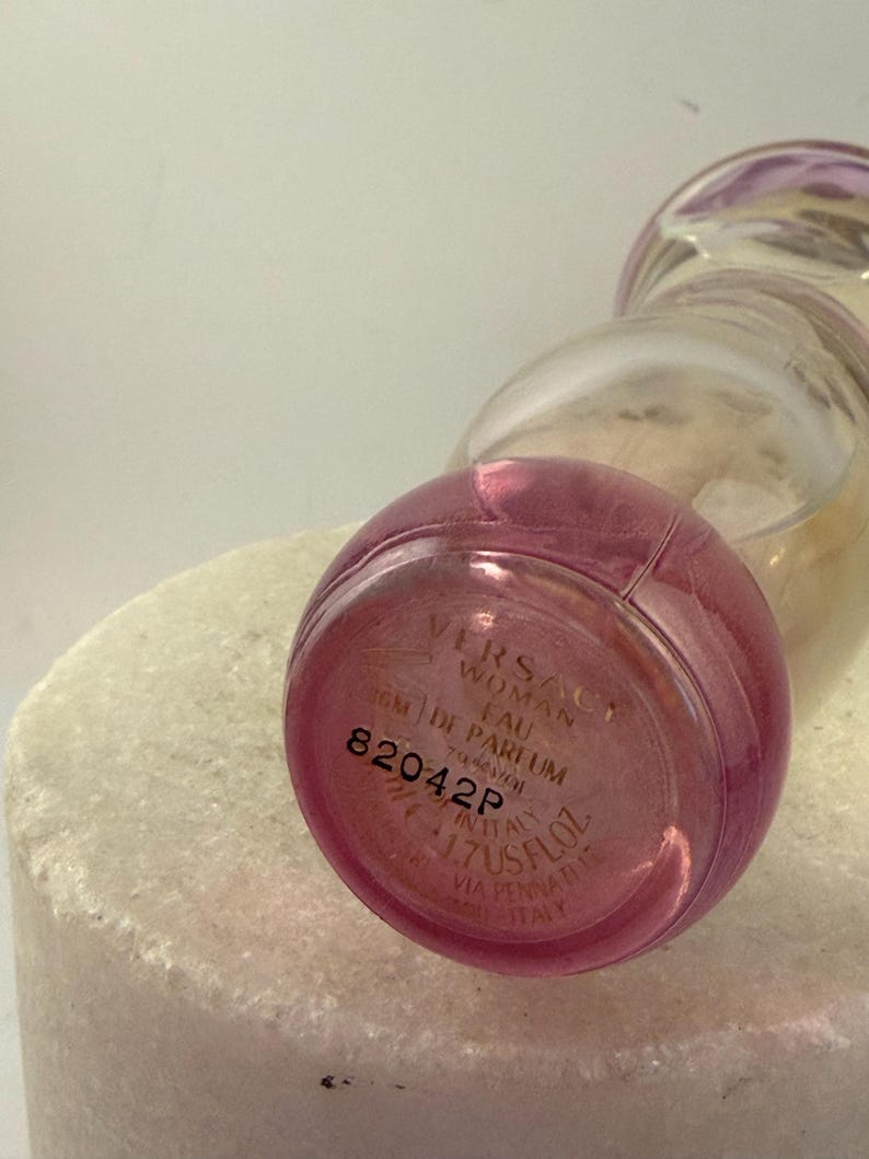 Puede incluir: Un frasco de perfume de vidrio transparente con una base rosa y una parte superior redondeada. El frasco tiene el texto "VERSACE WOMAN EAU DE PARFUM" y "1.7 US FL.OZ." impreso en la parte inferior. El frasco est&aacute; sobre una superficie blanca.