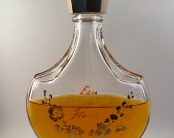 May include: A vintage perfume bottle with a clear glass body and a dark, geometric stopper. The bottle contains a golden-colored liquid and features the words "Eau de Fleurs" in elegant script, along with floral designs. The bottle is displayed on a white pedestal.