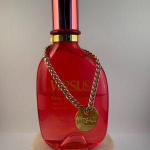 Könnte beinhalten: Eine rote Glasflasche mit Versus Versace Time For Dreamers Eau de Toilette Spray. Die Flasche hat eine goldene Kette um den Hals und ein goldenes Etikett mit dem Versace-Logo. Die Flasche ist 125 ml.
