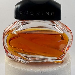 May include: A small perfume bottle with a clear glass body and a black cap. The bottle contains a golden-brown liquid and is labelled "KNOWING" on the cap. The bottle sits on a white, textured surface.