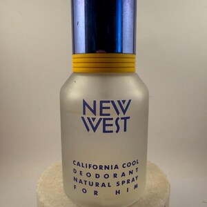Könnte beinhalten: Eine Flasche Deodorant "NEW WEST" aus mattiertem Glas. Die Flasche hat einen blauen Metallverschluss und ein gelbes Band. Der Text "CALIFORNIA COOL DEODORANT NATURAL SPRAY FOR HIM" ist auf der Flasche aufgedruckt.