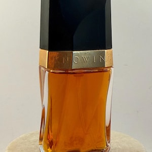 May include: A perfume bottle with a black cap and a gold band that reads "KNOWING". The bottle is filled with a golden-brown liquid and has a clear, twisted glass design. The bottle is displayed on a white, round surface.