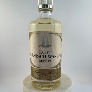 May include: A clear glass bottle with a gold label and a gold cap. The label reads "Echt K&ouml;lnisch Wasser" and "Doppelt". The bottle is sitting on a white surface.