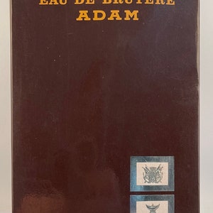 May include: A brown box with gold lettering that reads "EAU DE BRUYERE ADAM". The box has three silver stickers with images of a crest, a vase, and a leaf.