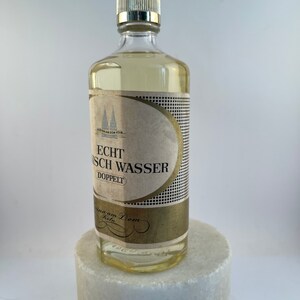 May include: A clear glass bottle with a gold label that reads "Echt K&ouml;lnisch Wasser Doppelt" and a gold cap. The bottle is sitting on a white marble surface.