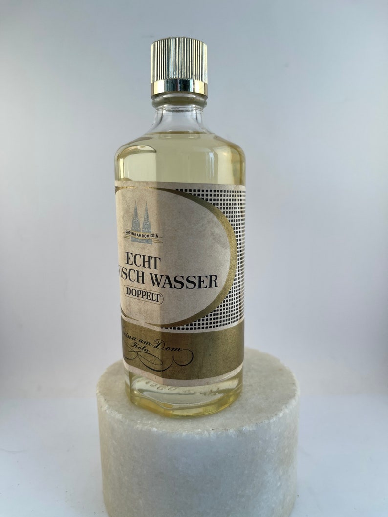 May include: A clear glass bottle with a gold label that reads "Echt K&ouml;lnisch Wasser Doppelt" and a gold cap. The bottle is sitting on a white marble surface.