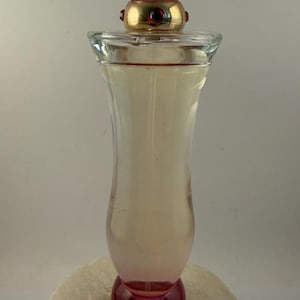 Puede incluir: Un frasco de perfume de vidrio transparente con una tapa rosa y dorada. El frasco tiene una base redondeada y un cuerpo delgado y curvado. La tapa es una gema rosa facetada. El perfume en su interior es de color rosa claro.