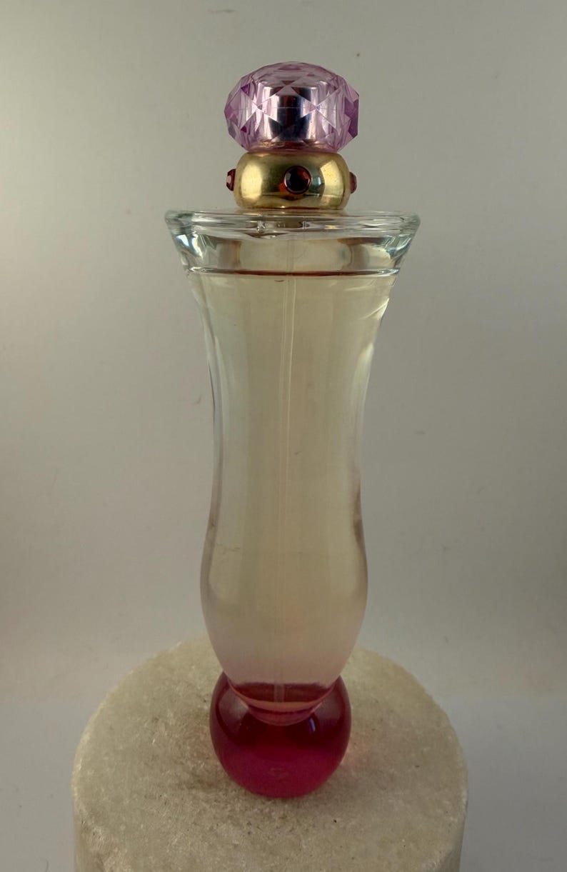 Puede incluir: Un frasco de perfume de vidrio transparente con una tapa rosa y dorada. El frasco tiene una base redondeada y un cuerpo delgado y curvado. La tapa es una gema rosa facetada. El perfume en su interior es de color rosa claro.