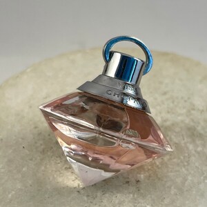Peut inclure: Un petit flacon de parfum en forme de diamant, transparent, avec un bouchon argenté et un anneau argenté. Le flacon est rempli d'un liquide rose clair. Le flacon porte l'inscription "Wish Chopard" sur le côté.