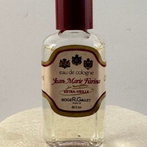 Op de afbeelding: Een klein, doorzichtig glazen flesje met Jean Marie Farina eau de cologne met een rode dop. Het etiket is geel met zwarte tekst en bevat drie wapenschilden. De tekst op het etiket luidt "eau de cologne Jean Marie Farina Extra-Vieille Roger & Gallet Paris 86% Vol."