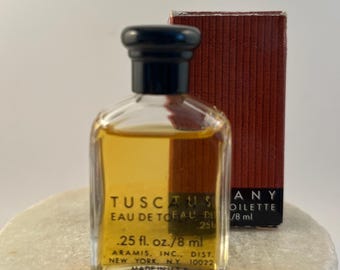 May include: A small glass bottle of Tuscany Eau de Toilette with a black cap, containing .25 fl. oz. of amber liquid. The label reads Aramis, Inc., Dist. New York, N.Y. 10022, Made in U.S.A. A matching rectangular box stands behind the bottle.