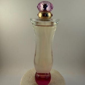 Puede incluir: Un frasco de perfume de vidrio transparente con una tapa enjoyada rosa y morada. La botella tiene una base redondeada y un efecto degradado rosa. El frasco de perfume est&aacute; sobre una superficie blanca texturizada.