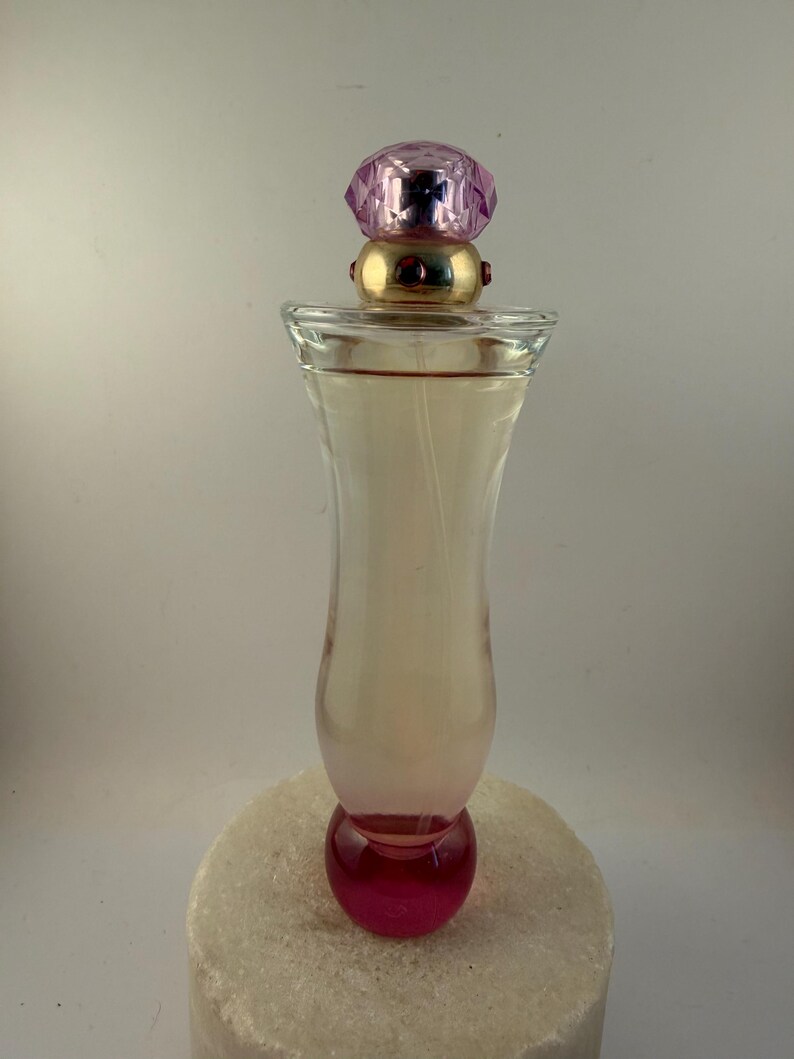 Puede incluir: Un frasco de perfume de vidrio transparente con una tapa enjoyada rosa y morada. La botella tiene una base redondeada y un efecto degradado rosa. El frasco de perfume est&aacute; sobre una superficie blanca texturizada.