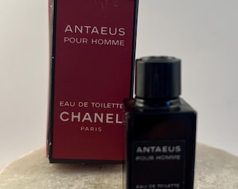 May include: A miniature bottle of Antaeus Pour Homme Eau de Toilette by Chanel with a black cap and label, accompanied by its matching rectangular box featuring a deep red front panel with white text reading Antaeus Pour Homme Chanel Paris.