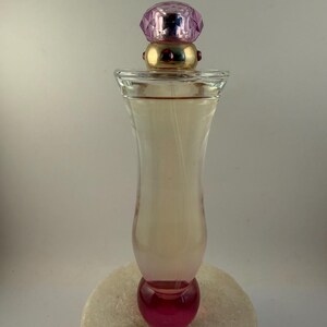 Puede incluir: Un frasco de perfume de vidrio transparente con una tapa rosa y dorada. La botella tiene una base redondeada y un efecto degradado rosa. La tapa es una gema rosa facetada. El frasco de perfume est&aacute; sobre una superficie blanca.