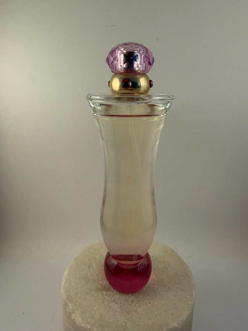 Puede incluir: Un frasco de perfume de vidrio transparente con una tapa rosa y dorada. La botella tiene una base redondeada y un efecto degradado rosa. La tapa es una gema rosa facetada. El frasco de perfume est&aacute; sobre una superficie blanca.