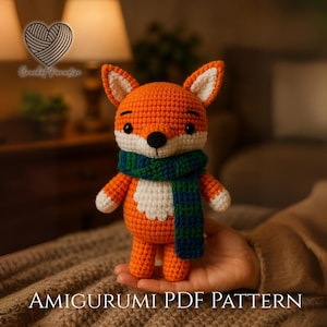 Op de afbeelding: Een gehaakt amigurumi vossenspeeltje van oranje en wit garen, met zwarte ogen en een neus, met een donkerblauwe en groene gestreepte sjaal. De tekst "Crochet Paradise" en "AMIGURUMI PDF PATTERN" is zichtbaar op de afbeelding.
