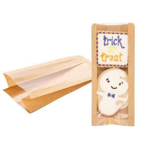May include: Two brown paper bags with clear plastic windows. One bag is lying flat, while the other stands upright containing two decorated cookies. The top cookie is square with the text trick or treat, and the bottom cookie is a white ghost.