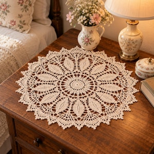May include: A round, cream-colored crochet doily featuring an intricate, repeating leaf-like pattern radiating from a central circular motif. The edges are finished with a delicate, scalloped lace border, creating a detailed, circular textile design.