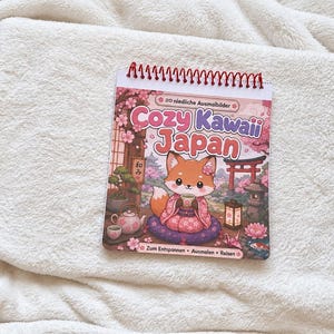 Könnte beinhalten: Ein spiralgebundenes Malbuch mit dem Titel Cozy Kawaii Japan, das einen Cartoon-Fuchs im Kimono mit einer Teetasse zeigt. Das Cover zeigt Kirschblüten, ein rotes Torii, eine Steinlaterne und eine Teekanne. Der Text lautet 20 niedliche Ausmalbilder und Zum Entspannen Ausmalen Reisen.