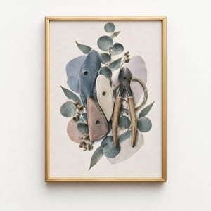 May include: A framed print featuring a composition of three ceramic pottery ribs in blue, beige, and mauve, alongside a pair of metal wire cutters, a coil of wire, and sprigs of eucalyptus leaves with small round seed pods on a light background.