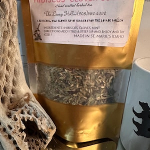 May include: A gold-colored stand-up pouch containing herbal tea, labeled Hibiscus Clover Cooler. The label lists ingredients as hibiscus, cloves, and mint, with instructions to add 1 tablespoon, steep, and enjoy, made in St. Marie's, Idaho.