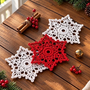 May include: Three crocheted snowflake ornaments, two white and one red, featuring intricate lace patterns with a central circular opening. The snowflakes are arranged on a wooden surface alongside cinnamon sticks, red berries, and a gold bell.