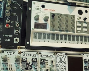 以下が含まれることがあります： モジュラーシンセサイザーのセットアップのクローズアップ。画像には、Korg Volca Sample、VCOモジュール、および「Envelope」、「VCF」、「Multiples」、「Delay」とラベル付けされたその他のコンポーネントを含む、さまざまな電子音楽モジュールが表示されています。モジュールはラックにマウントされています。