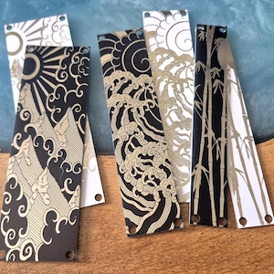 May include: A collection of decorative bookmarks with Asian-inspired designs. The bookmarks feature black and white backgrounds with gold accents. Designs include mountains, sun, waves, clouds, and bamboo. Each bookmark has a hole at the top.