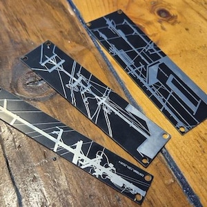 Puede incluir: Tres piezas rectangulares de metal negro con un diseño plateado de líneas eléctricas y postes de servicios públicos. Las piezas están dispuestas sobre una superficie de madera. Cada pieza tiene un pequeño agujero en cada extremo.