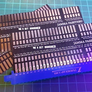 May include: Three black circuit boards with white text that reads "The 8 Bit Manshed" and "JAMMA PASSTHRU". A blue connector with white text that reads "JAMMA 56P-3.965" is in the foreground.